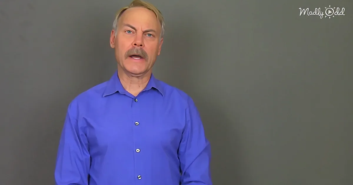 A man in a blue shirt stands against a gray background, looking directly at the camera.