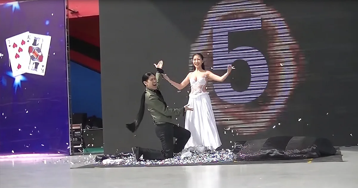 A woman in a white dress stands on stage with a man kneeling, surrounded by confetti and a large "5" backdrop.