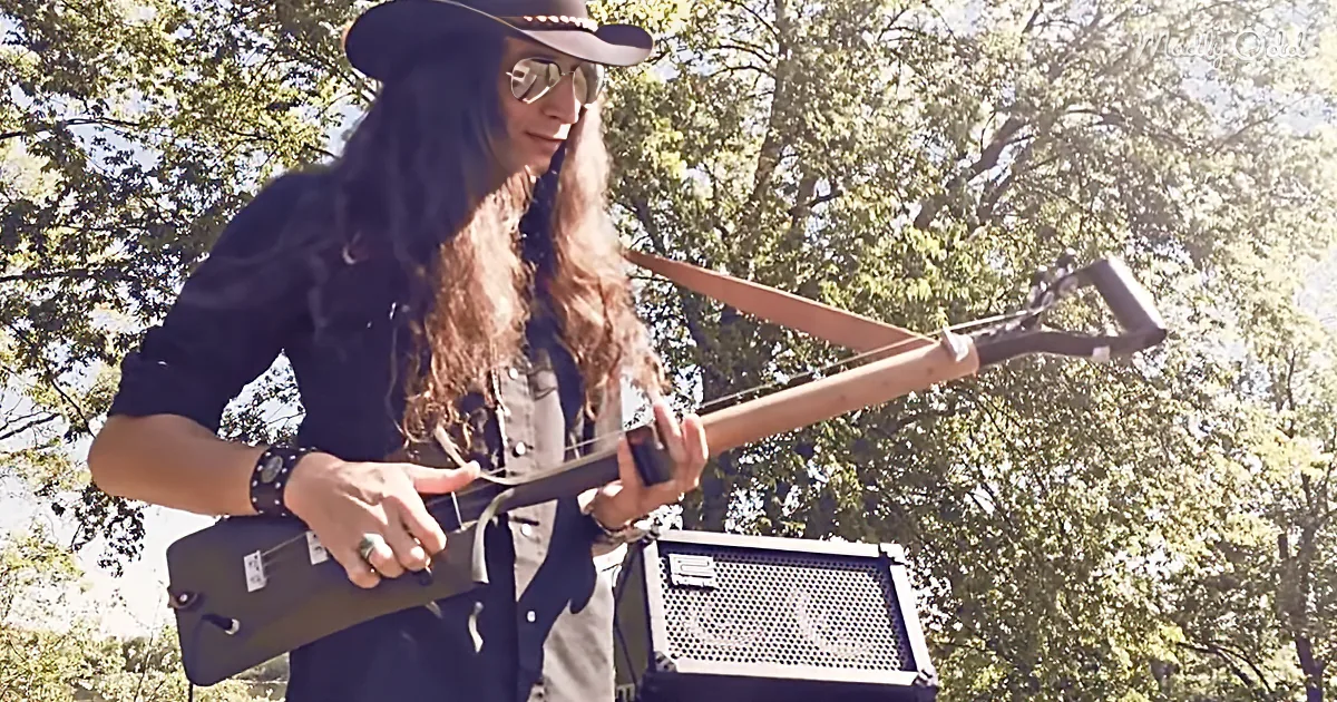 A person with long hair and a hat plays a guitar-like instrument outdoors, with trees and sunlight in the background.