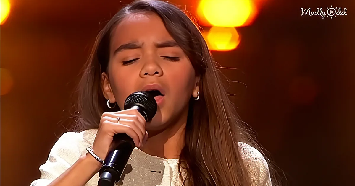 A young woman with long hair sings passionately into a microphone, her eyes closed, in a warmly lit setting.