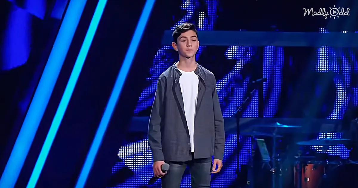 A young man stands on stage, holding a microphone, wearing a white shirt and dark jacket, with a blue-lit backdrop and...