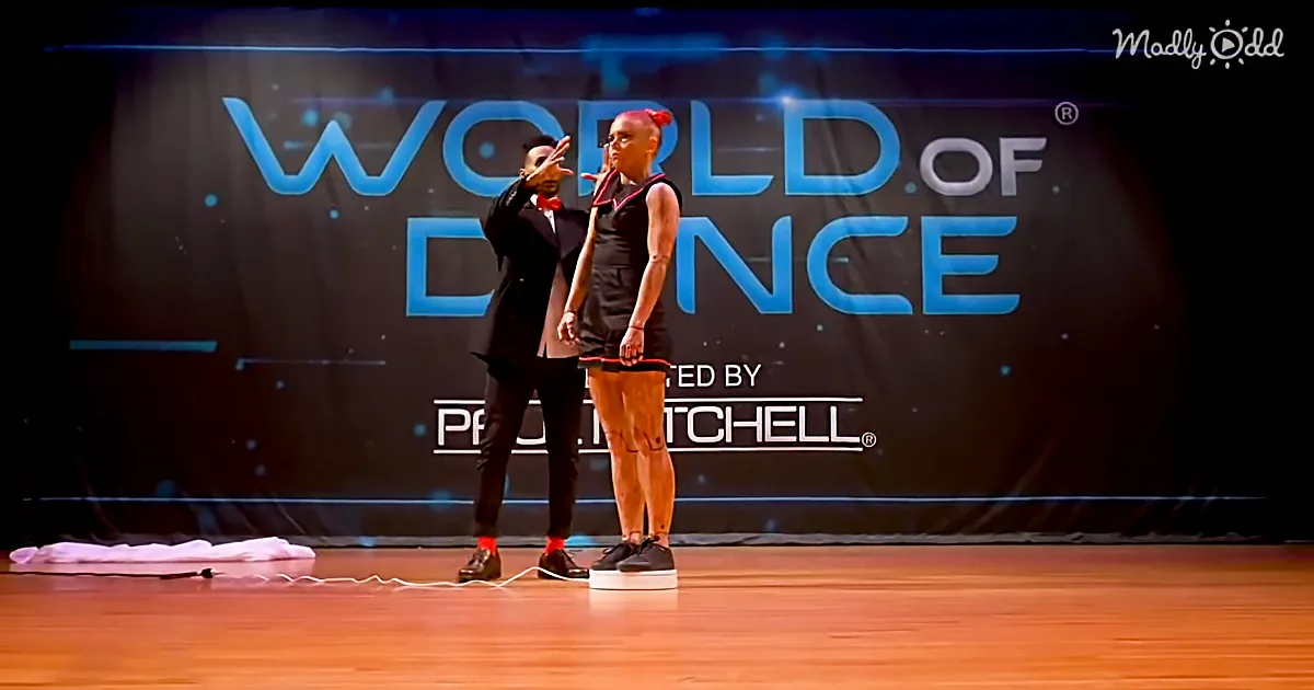 A man and woman stand on stage at the World of Dance event, with a backdrop featuring the show's name and sponsor.