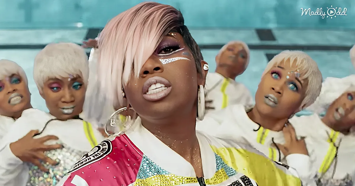 A person with pink and white hair, wearing a colorful outfit, stands in front of a group of people in white shirts with...
