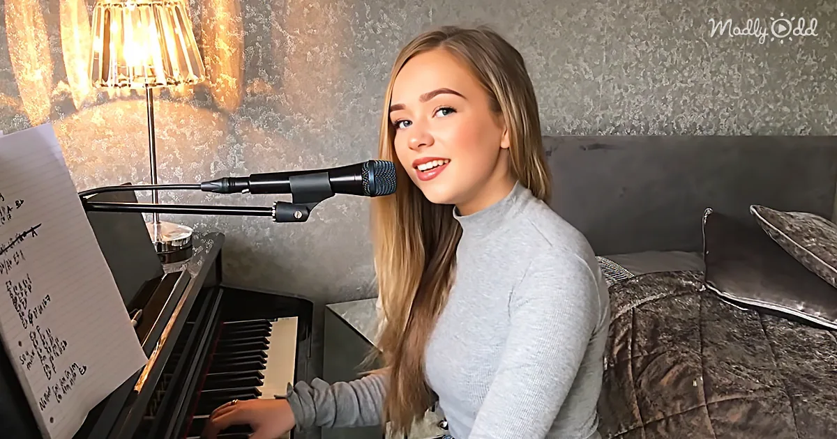 A person with long hair sits at a piano, singing into a microphone, with a lamp and bed in the background.