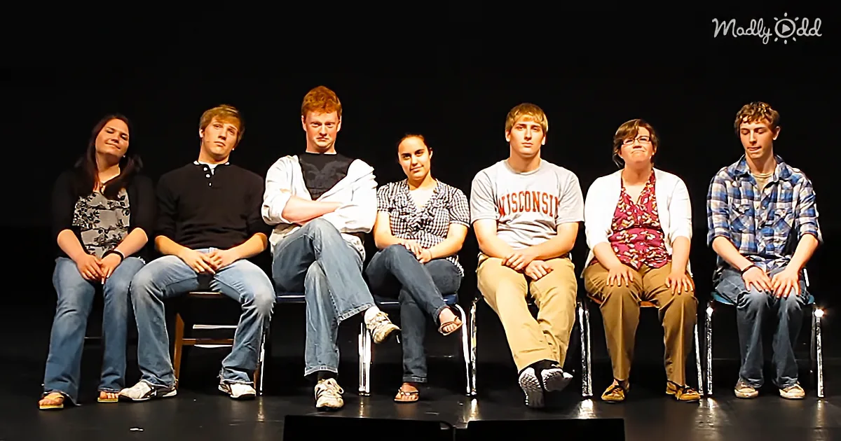 A group of eight people sit on stage chairs, some looking serious, others relaxed, in a dimly lit setting.