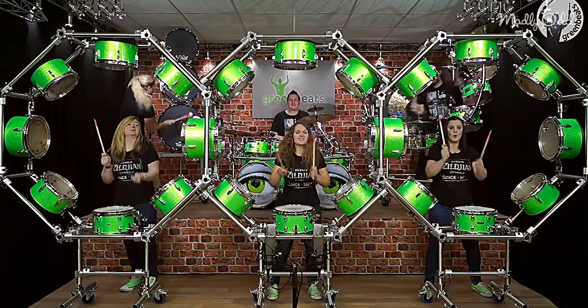 A person plays a drum set surrounded by multiple green drum kits in a studio setting.