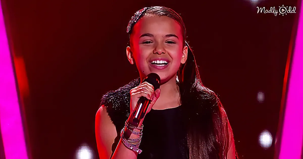 A young girl smiles while holding a microphone, wearing a black outfit and bracelets, with a red and pink background.