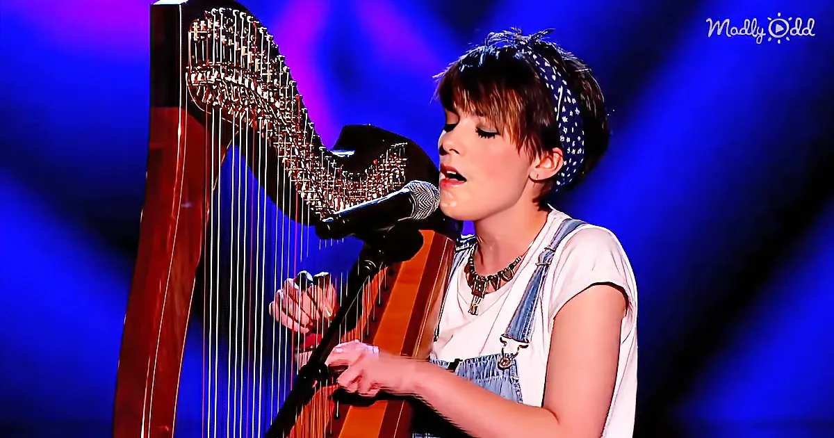 A person plays a harp and sings into a microphone on stage with a blue background.