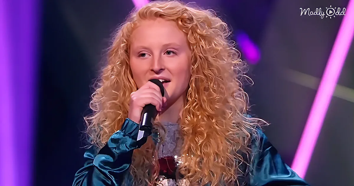 A person with long, curly blonde hair holds a microphone, wearing a teal outfit, against a purple-lit background.