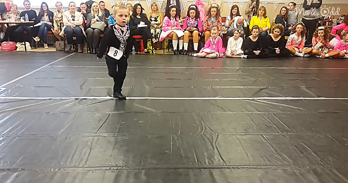 A young boy in a black outfit with a number 8 on his chest stands on a stage, surrounded by seated spectators.