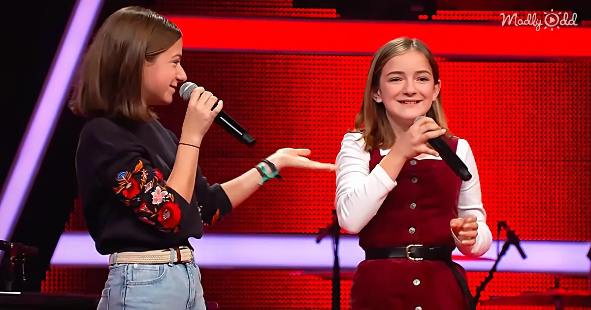 Two girls on stage, one in jeans and black shirt, the other in a red dress, both holding microphones and smiling.
