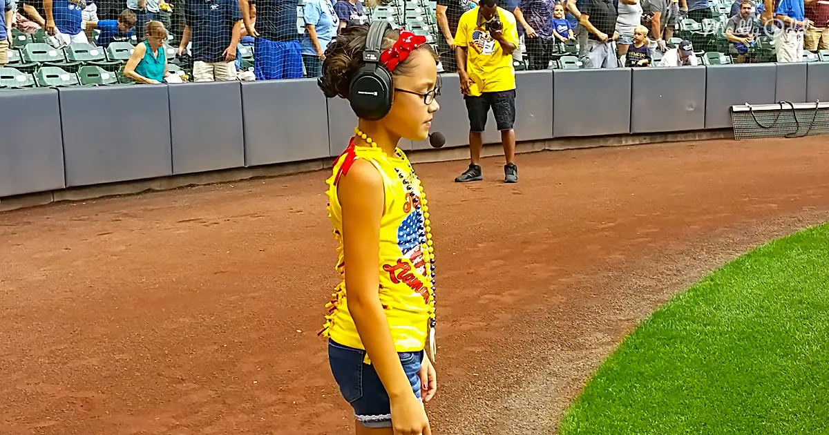 A young girl wearing a yellow shirt, headphones, and a red bow stands on a baseball field, facing away from the camera.