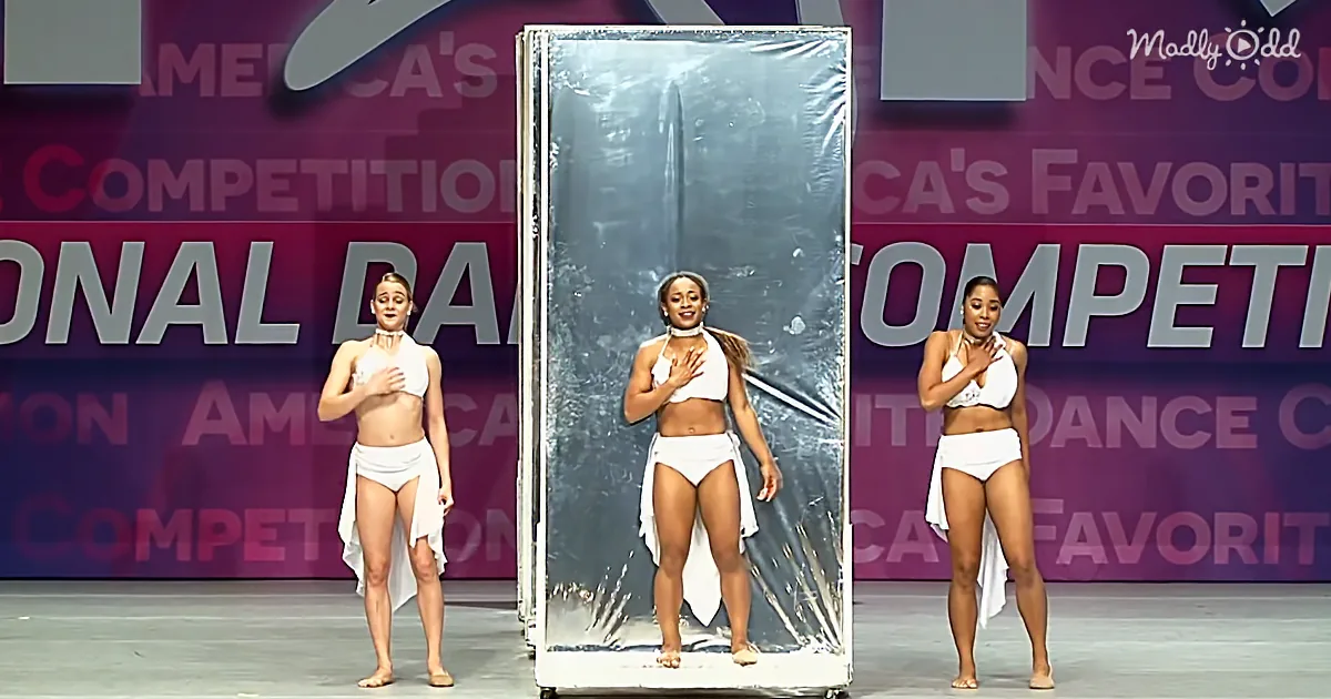 A woman in a white bikini and white scarf stands on stage, hands on chest, with a reflective backdrop behind her.