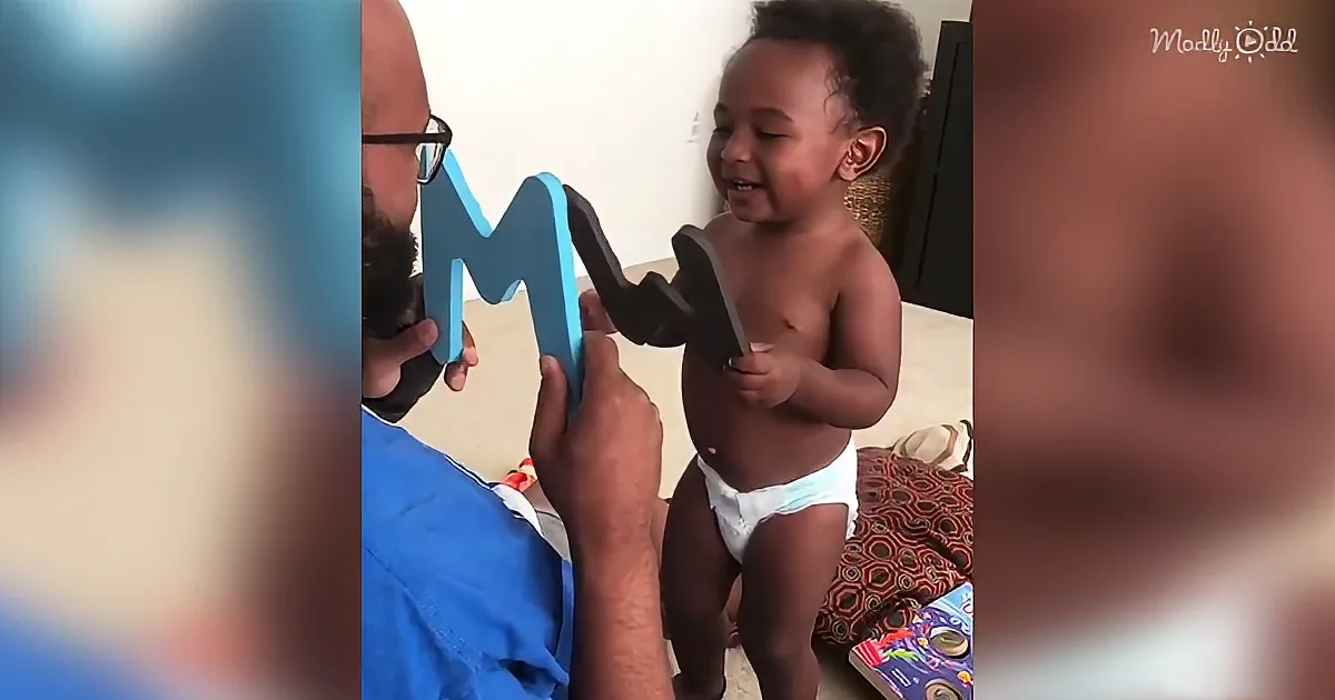 A baby in a diaper holds a letter "M" while a person in a blue shirt holds another part of the letter.