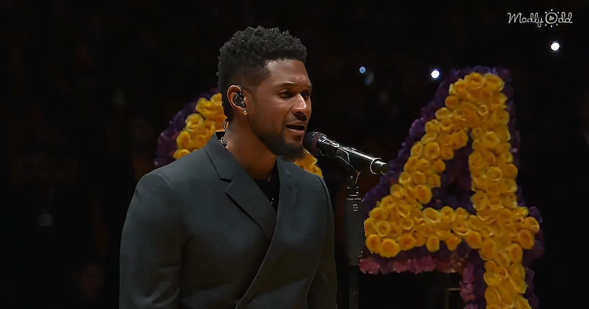 A man in a suit sings into a microphone, wearing earbuds, with a floral display in the background.
