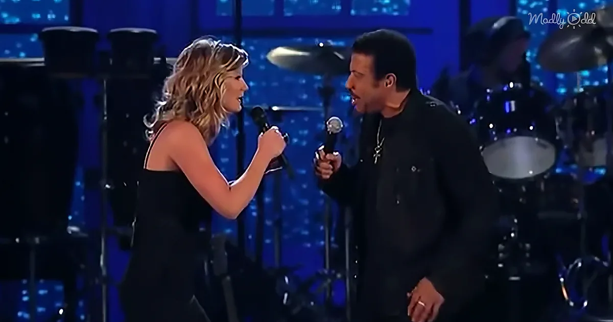 A woman and man sing together on stage, both holding microphones, with a band in the background.