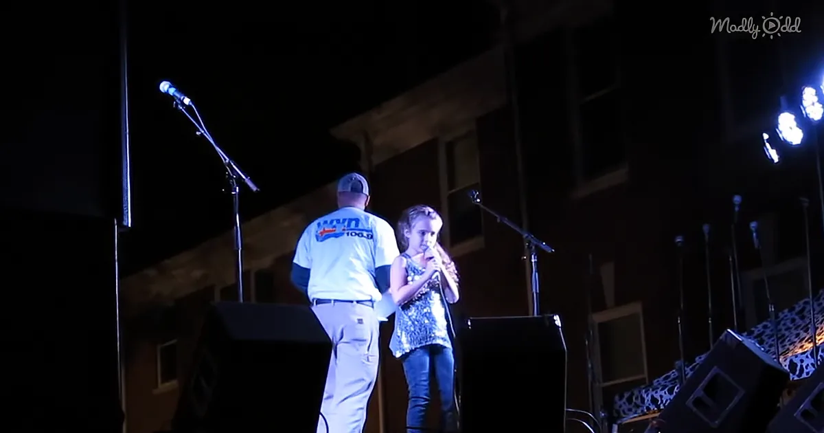 A young girl sings into a microphone on stage with a man in a white shirt.