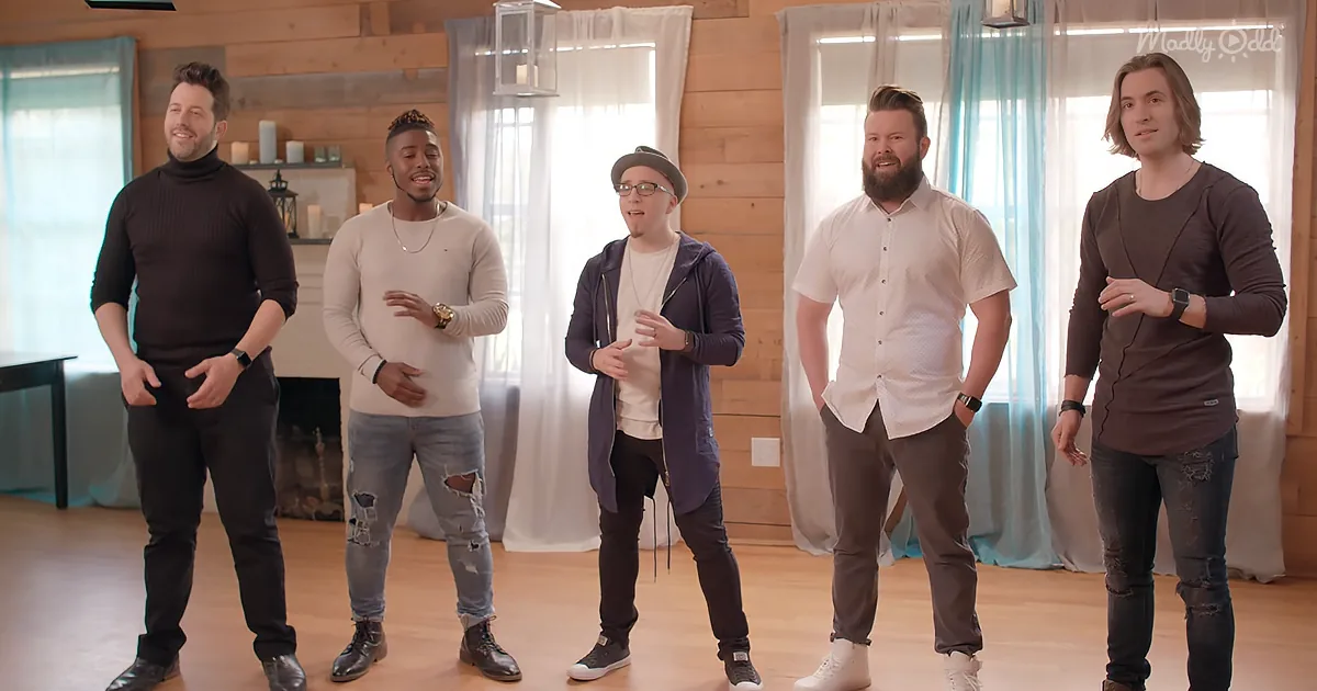 Five men in casual attire stand in a room with wooden walls and windows, appearing to sing or speak.