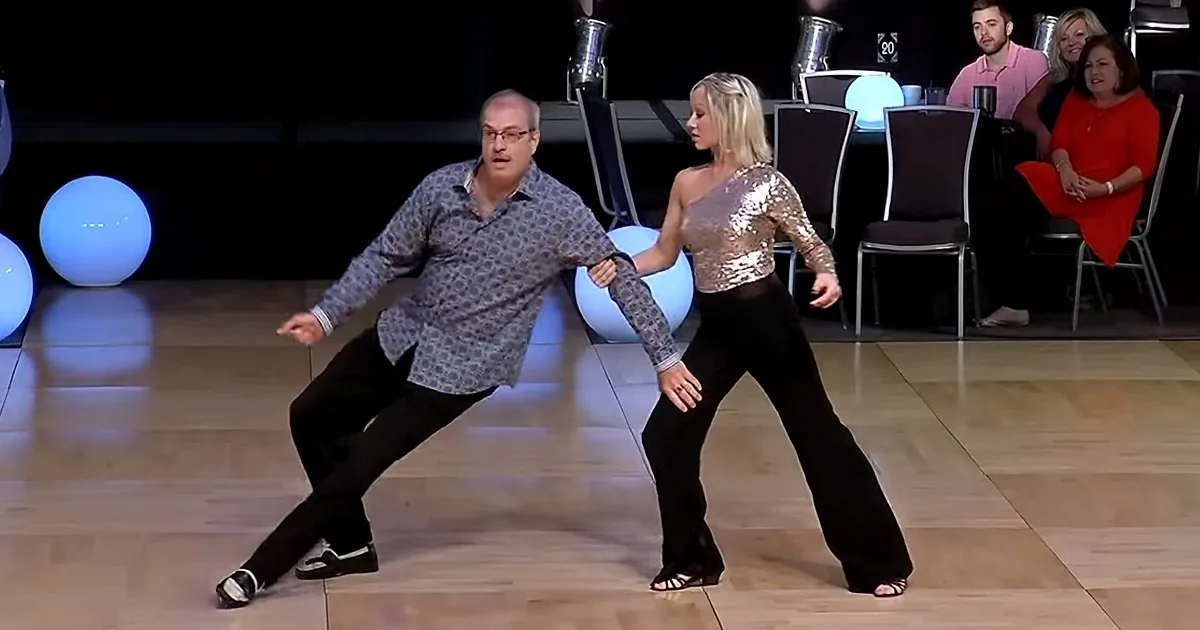 A man and woman are dancing on a stage with blue spheres in the background, surrounded by an audience.