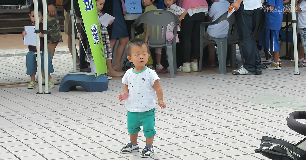 A toddler in a white shirt and green pants stands on a tiled floor, surrounded by adults and children.