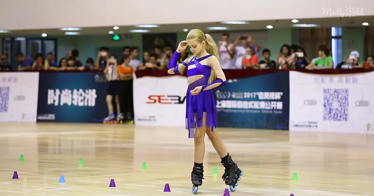 A young girl in a purple outfit rollerblades on a wooden floor with cones, surrounded by spectators.