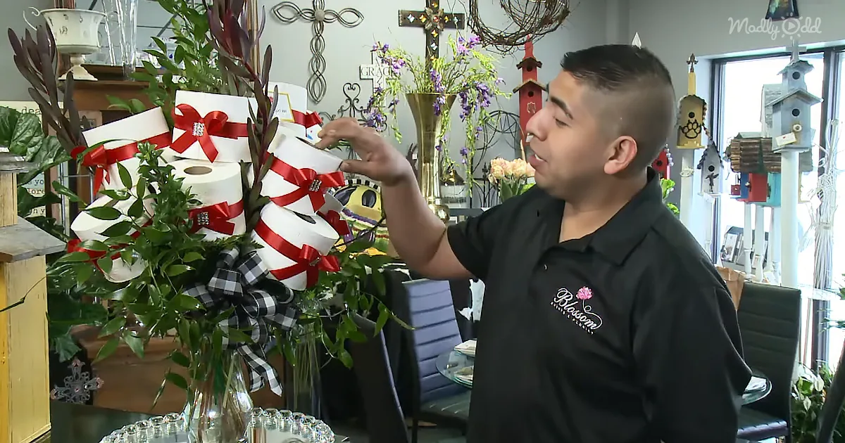 A man in a black shirt with "Blossom" logo stands beside a decorative wreath with toilet paper rolls and red bows.