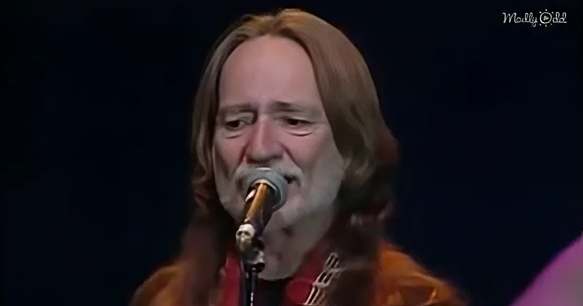 A person with long hair and a beard sings into a microphone, wearing an orange shirt.