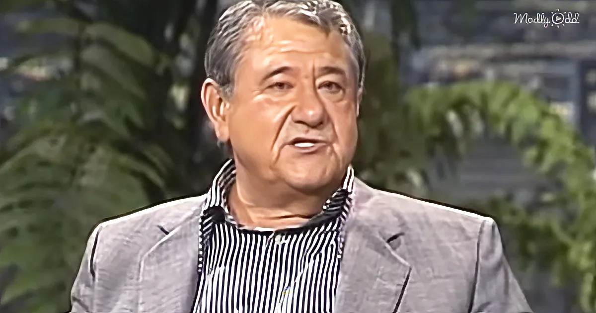 A man in a striped shirt and gray blazer is speaking, with green foliage in the background.