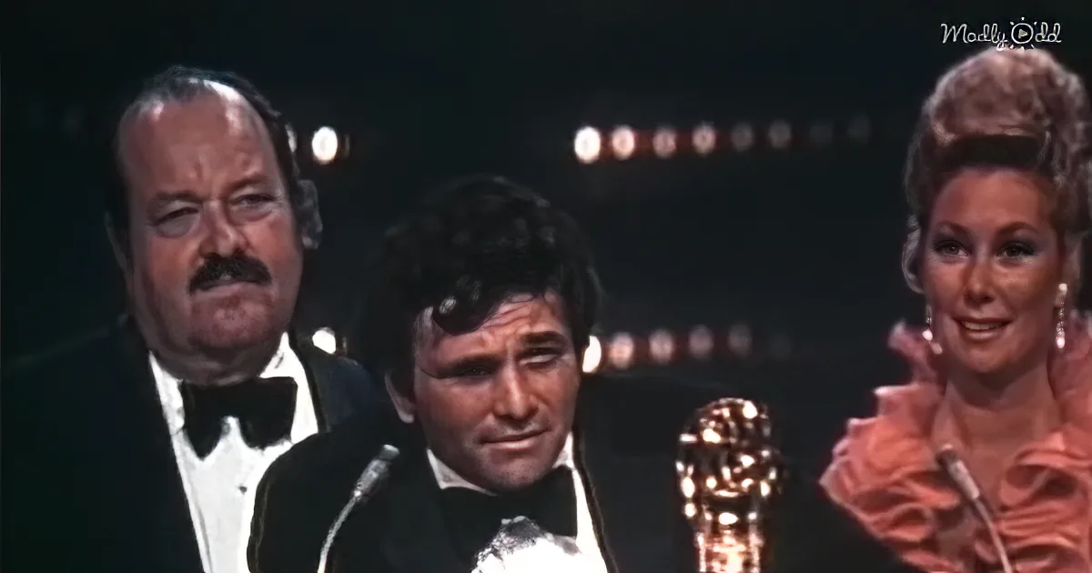 A man in a tuxedo speaks into a microphone, holding an Oscar statue, with two others in formal attire behind him.