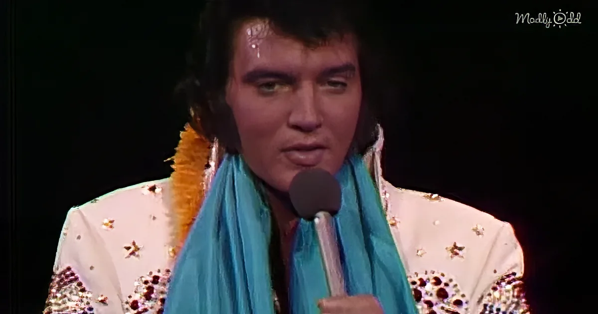 Elvis Presley in a white sequined suit with a blue scarf, holding a microphone.