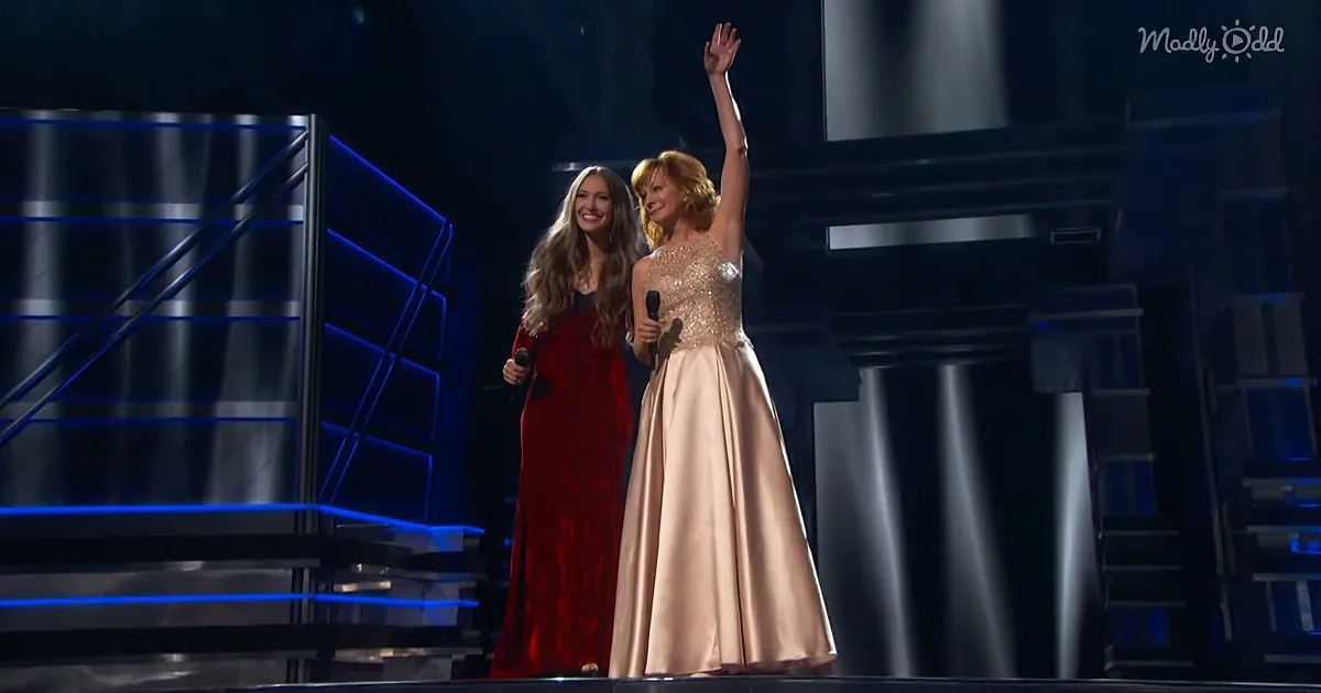 Two women in elegant dresses on stage, one raising her arm, both holding microphones.