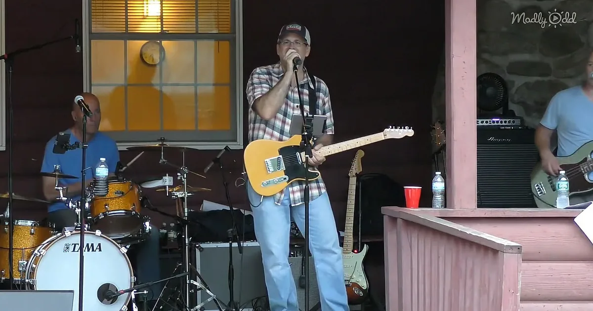 A man in a plaid shirt plays a yellow guitar and sings into a microphone.