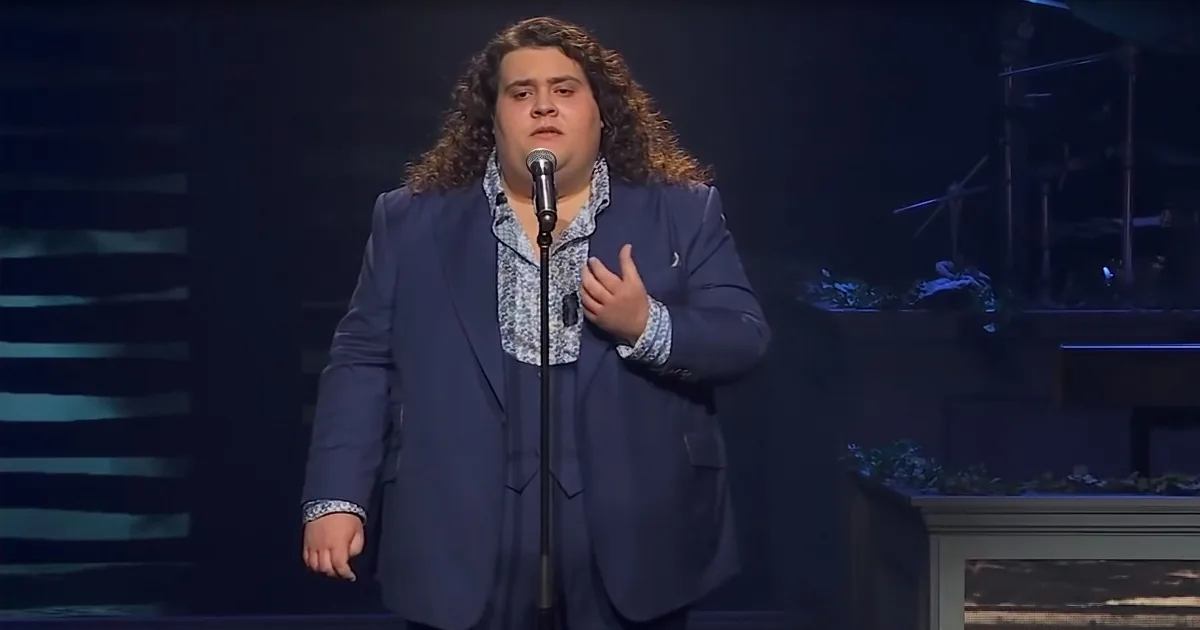 A man in a blue suit stands on stage, holding a microphone, with his hand near his chest.