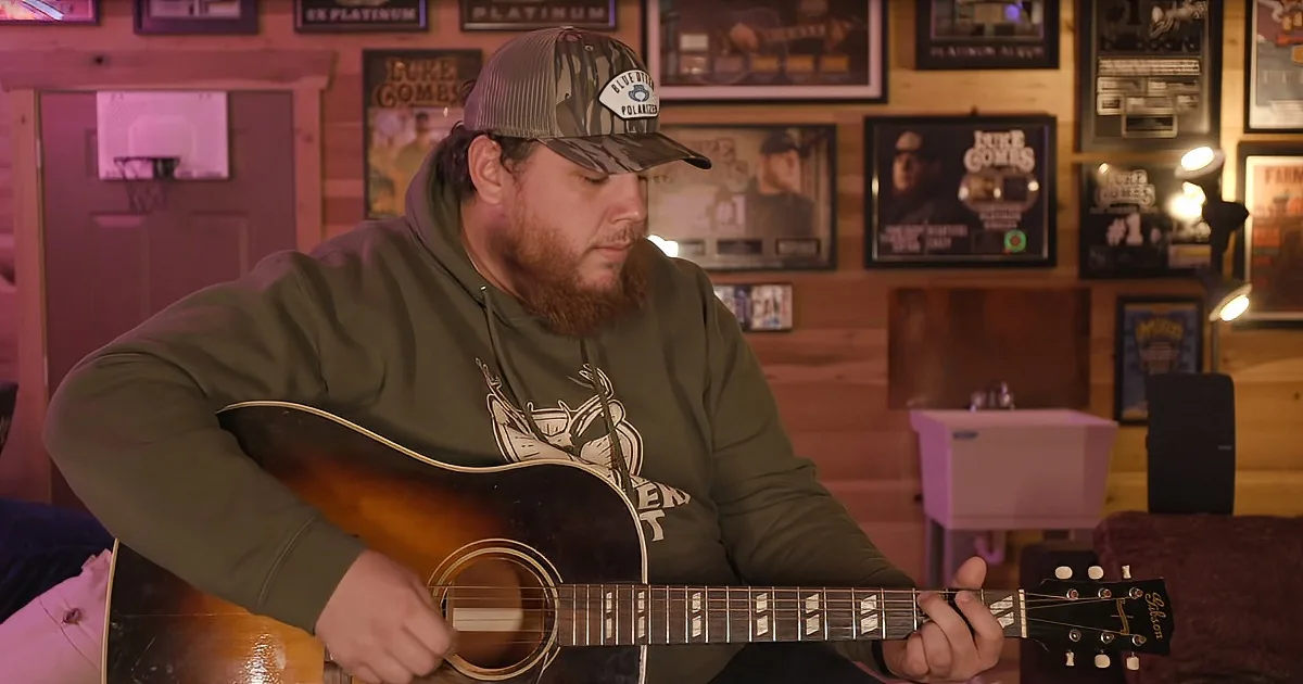 A man with a beard and a cap plays an acoustic guitar in a room adorned with framed music awards and posters.