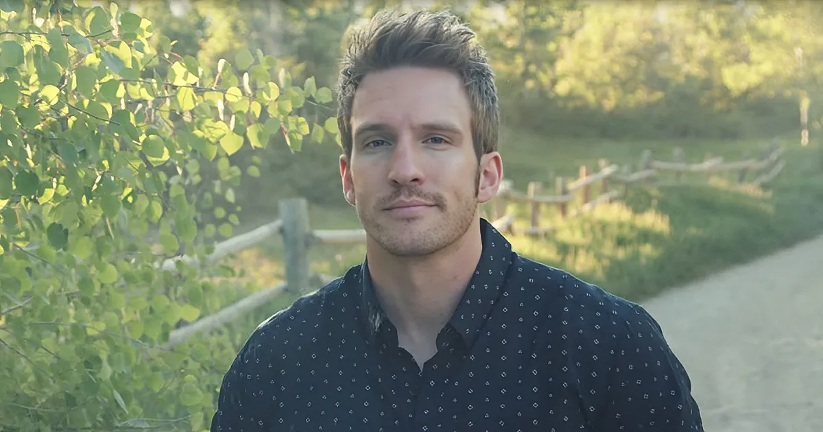 A man with short brown hair and a black patterned shirt stands outdoors, with greenery and a wooden fence in the background.