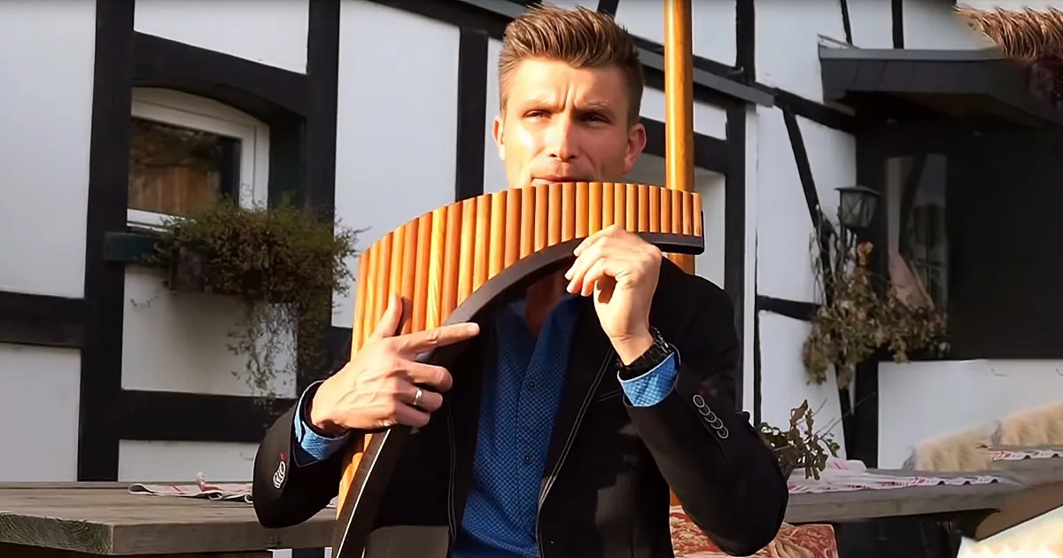 A man in a suit holds a wooden pan flute, partially covering his face, in front of a half-timbered house.
