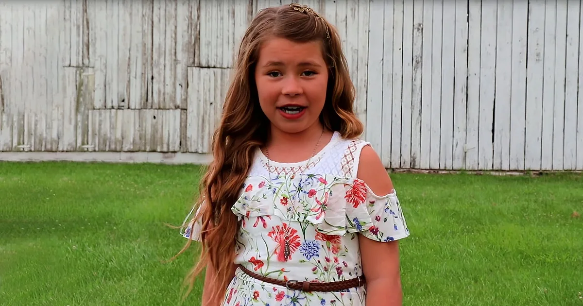 A young girl with long brown hair, wearing a floral dress, stands in front of a white wooden fence on a grassy field.