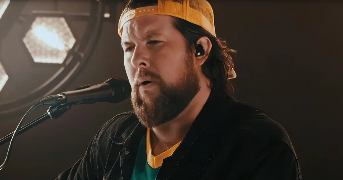 A man with a beard and a yellow cap sings into a microphone, wearing a black jacket and a green shirt.