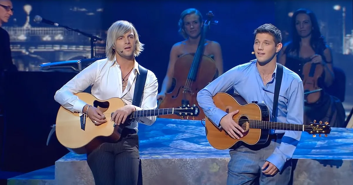 Two men play acoustic guitars on stage, one in white, the other in blue, with a cello player in the background.