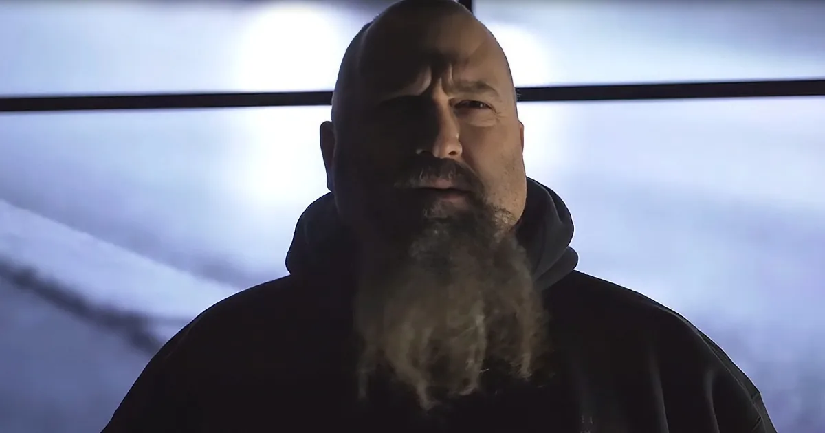 A man with a long beard and mustache, wearing a dark hoodie, stands in front of a snowy landscape with a bright light in...