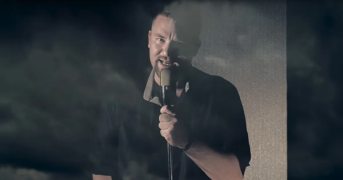 A man in a dark suit holds a microphone, singing against a smoky, textured background.
