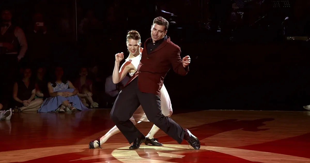 A couple in formal attire dances gracefully on a brightly lit stage, surrounded by an audience.