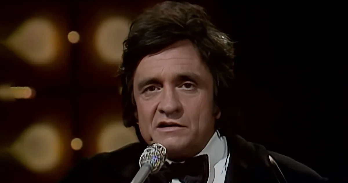 A man in a tuxedo sings into a microphone, with blurred lights in the background.