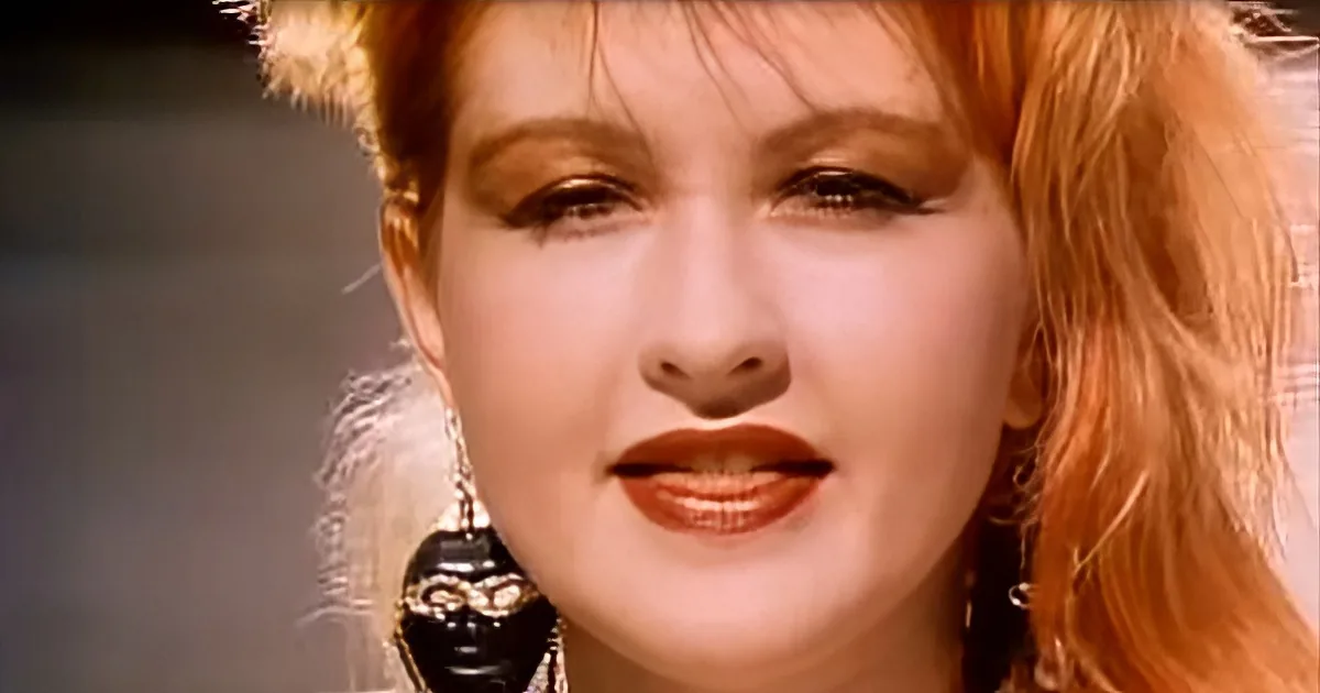 A woman with red hair and large earrings is shown in a close-up, wearing makeup and a fur-like headband.