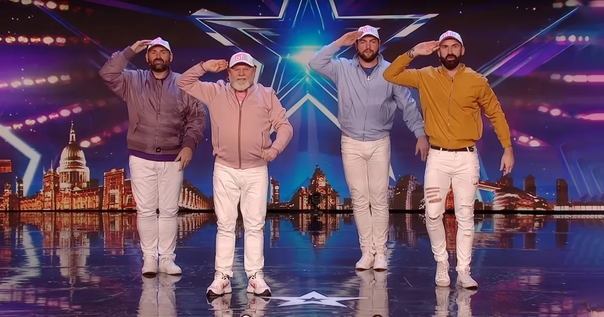 Four men in white pants and caps salute on a stage with a cityscape backdrop.