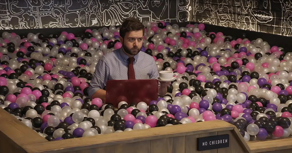 A man sits at a desk surrounded by colorful balls, working on a laptop with a coffee cup nearby.