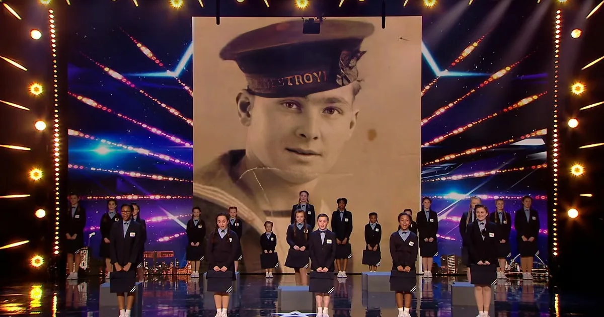 A group of children in school uniforms stands in front of a large screen showing a man in a military uniform.
