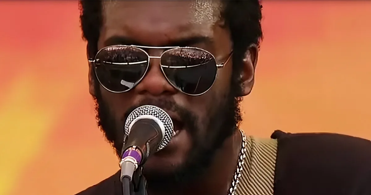 A person wearing aviator sunglasses and a necklace sings into a microphone against a blurred orange background.