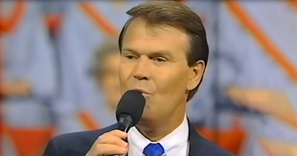 A man in a suit and blue tie speaks into a microphone, holding it with his right hand.