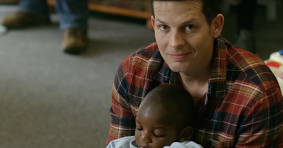 A man in a plaid shirt holds a baby, both looking down, in a casual indoor setting.
